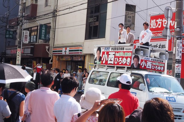 強い日差しの中、大勢の人が足を止めて演説を聞く。司会は村越祐民千葉５区総支部長。